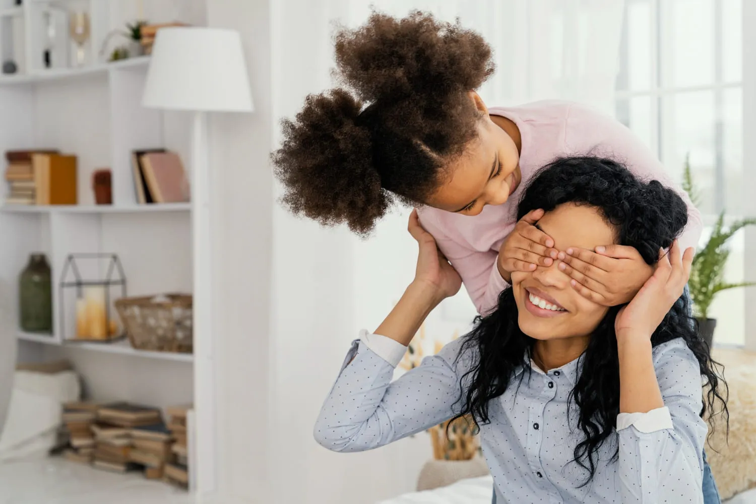 A young girl playfully covering her mother's eyes at home, both laughing