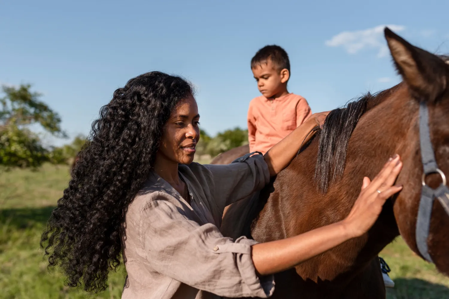 A mother with long curly hair stroking a horse while her small son sits on its back