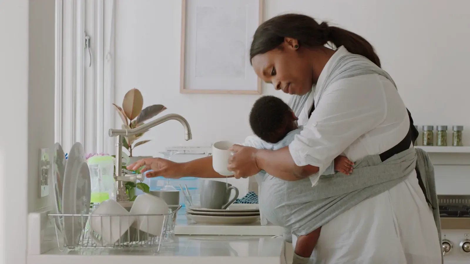 A mother carrying her baby in a sling while filling a cup at the kitchen sink